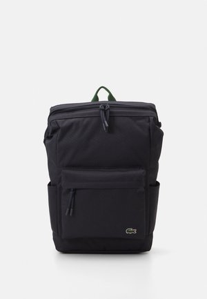 Črni nahrbtnik, izdelan iz trpežne tkanine, ima zgornje zapiranje na zadrgo, sprednji žep, ročaj v zeleni barvi in majhen logo.