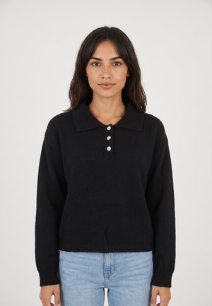 Donna con capelli scuri che indossa un maglione nero a maniche lunghe con colletto e tre bottoni bianchi, e jeans azzurri chiari, in piedi davanti a uno sfondo bianco.