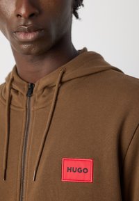 Tuvo atainis, kurā redzams cilvēks brūnā ar rāvējslēdzēju aizveramā kapucē ar sarkanu taisnstūra "HUGO" logo uz krūtīm.
