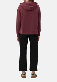 Persona vestida con una sudadera burdeos, jeans cortos negros y zapatillas beige con detalles metálicos, de pie de espaldas.