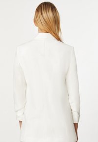 Biała marynarka o dopasowanym kroju z długimi rękawami, wyciętym kołnierzem i dopasowanym sylwetką. Posiada gładki materiał i minimalne detale szwów.