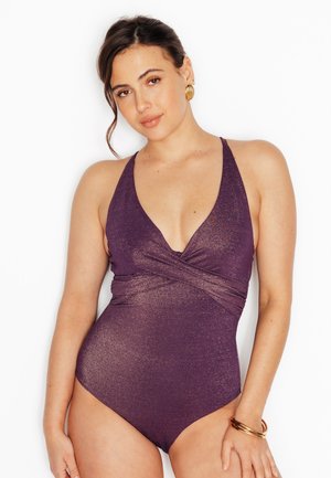Femme portant un maillot de bain mauve scintillant à col halter avec un détail froncé à la taille, des boucles d'oreilles anneaux dorés et des bracelets dorés sur fond blanc.