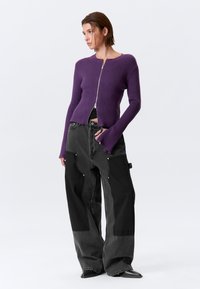 Cardigan violet en maille côtelée avec fermeture éclair, associé à un pantalon large noir fluide avec surpiqûres décoratives et garnitures métalliques.