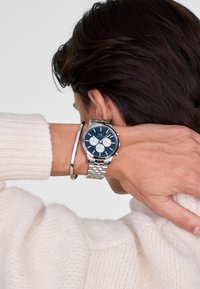 Orologio in acciaio inossidabile con quadrante blu, dettagli argentati, funzioni cronografiche e bracciale a rete argentato, abbinato a un sottile bracciale argentato.