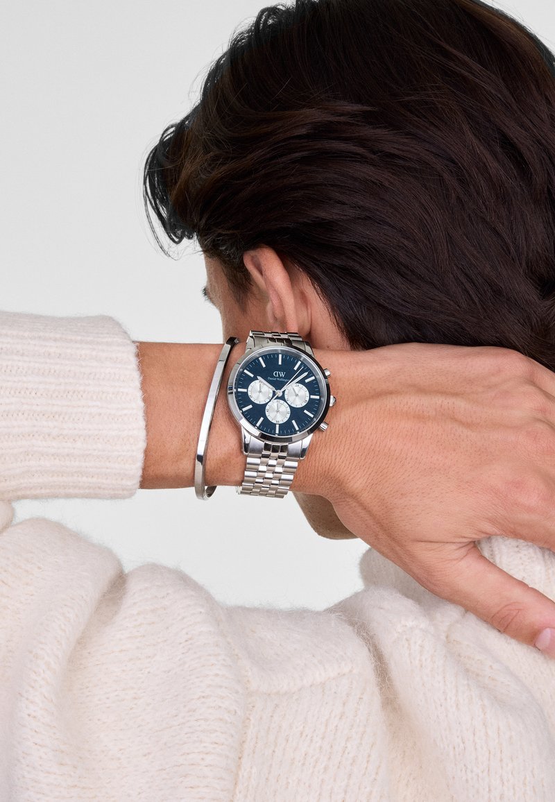 Orologio in acciaio inossidabile con quadrante blu, dettagli argentati, funzioni cronografiche e bracciale a rete argentato, abbinato a un sottile bracciale argentato.
