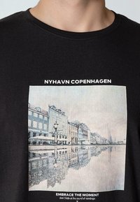 Černé bavlněné tričko se grafikou Nyhavnu, Kodaně, s barevnými budovami a odrazy ve vodě. Text nad a pod obrázkem.