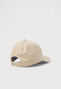 Beige színű baseball sapka, állítható snapback zárással, látható varrással és szellőző lyukakkal, egy fehér felületen pihen.