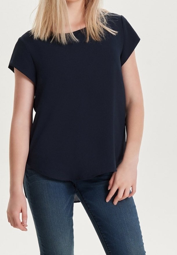 Femme portant un chemisier bleu marine uni à manches courtes et un jean bleu, debout avec une main posée sur la hanche.