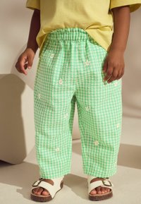 Grüne karierte Baumwollhose mit elastischem Bund, verziert mit weißen Gänseblümchen-Stickereien. Kombiniert mit weißen Sandalen und einem gelben Oberteil.