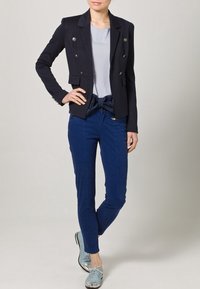 Marinblå kavaj med dubbla silverknappar, grå topp, blå figurnära byxor och ljusblå skor med metalliska accenter.