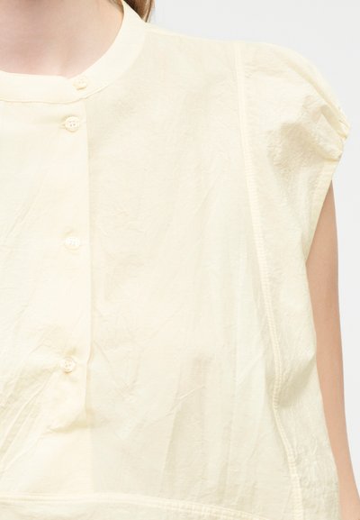 Κοντινό πλάνο ενός ατόμου που φοράει μια μπλούζα σε εκρού χρώμα με κοντά μανίκια, κουμπιά μπροστά και ελαφρώς φουσκωτή λεπτομέρεια στους ώμους.