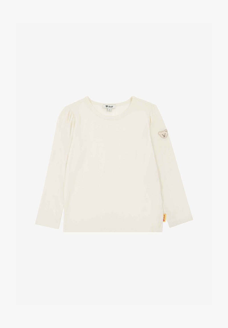 Chemise à manches longues pour tout-petit de couleur crème avec encolure ronde, petite applique en forme d'ourson sur la manche gauche, et étiquette de marque jaune à l'ourlet.
