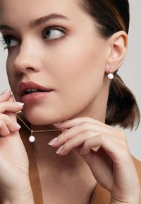 Złoty naszyjnik z białym okrągłym wisiorkiem oraz pasującymi kolczykami, oba z gładką powierzchnią, noszone przez osobę o jasnej skórze.