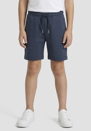 Enfant portant un short bleu marine à cordon de serrage et des baskets blanches, debout devant un fond clair uni.