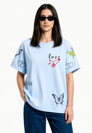 Femme aux cheveux foncés portant des lunettes de soleil noires, un t-shirt bleu clair avec le texte "love", illustrations d'épingle de sûreté, cœurs, papillon, fleur et pansement.