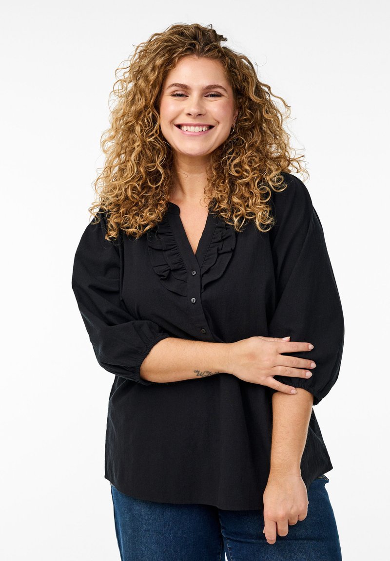 Schwarze Bluse mit Rüschenausschnitt, halblangen Ärmeln und Knopfverschluss. Weicher Stoff mit lockerer Passform, kombiniert mit blauen Jeans.