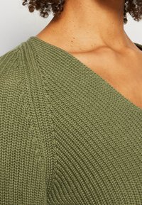 Pull en maille vert olive présentant un motif côtelé texturé, un col en V et une transition d'épaule sans couture. Vue en gros plan montrant les détails.