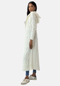 Weißer, gestrickter langer Cardigan mit Kapuze, ausgestattet mit Zopfstrickmustern und gerippten Bündchen, kombiniert mit blauen Jeans und weißen Turnschuhen.