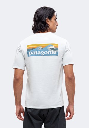 Mand med mørkt hår iført hvid Patagonia t-shirt med farverig bølgestreg på ryggen, parret med sorte bukser, med ryggen mod kameraet.