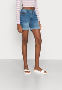 Hoikka henkilö, joka on pukeutunut ylös käännettyihin sinisiin farkkushortseihin, pitkätakkiin laventelin ja kermakerroksen raidallisessa lyhyessä paidassa sekä valkoisiin liukumäki-sandaaleihin.