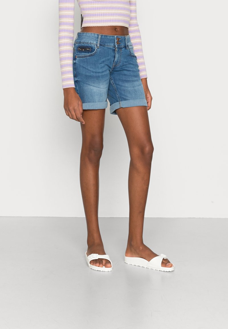 Hoikka henkilö, joka on pukeutunut ylös käännettyihin sinisiin farkkushortseihin, pitkätakkiin laventelin ja kermakerroksen raidallisessa lyhyessä paidassa sekä valkoisiin liukumäki-sandaaleihin.