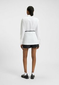 White long-sleeve shirt with a fitted silhouette, cinched at the waist by a black belt, paired with black shorts and black shoes.