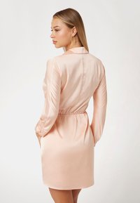 Satin persikinis suknelė su ilgomis rankovėmis, elastinga juosmens dalimi ir lygaus paviršiaus tekstūra. Yra apykaklė ir kelio ilgio apačia, rodoma iš nugaros.