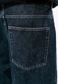 Jeans din denim închis cu un prim-plan al buzunarului din spate. Prezintă un contur cusut, o ușoară decolorare și un aspect texturat al țesăturii.