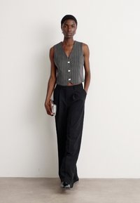 Woman wearing a sleeveless gray pinstripe vest, black wide-leg trousers, holding a silver clutch, walking against a plain white background.