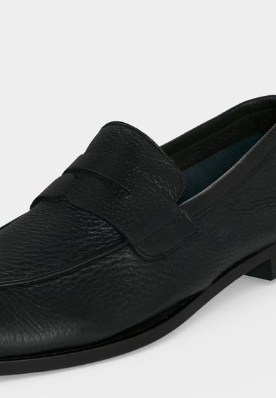 Mocassin homme en cuir noir avec une surface texturée et un détail de bride sur le dessus, présenté de profil sur fond blanc.