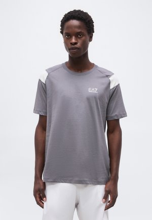 Νεαρός που φοράει γκρι μπλουζάκι EA7 με λευκές λεπτομέρειες στους ώμους και λευκό παντελόνι, στέκεται μπροστά σε απλό ανοιχτό φόντο.