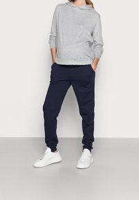 Šedá mikina s kapucí, námořnické tepláky s bočními kapsami, kombinované s bílými sportovními botami, vše na neutrálním pozadí.