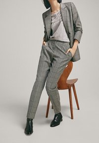 Costume à carreaux gris avec une veste cintrée et un pantalon fuselé, associé à un t-shirt gris. Bottines en cuir noir. Le mannequin est assis sur une chaise en bois.
