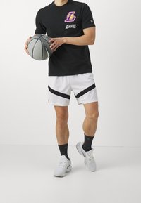 Svart t-shirt med kort ärm som har en lila och vit Los Angeles Lakers-logga, tillsammans med vita mesh-shorts med svarta accenter. Håller en basketboll.