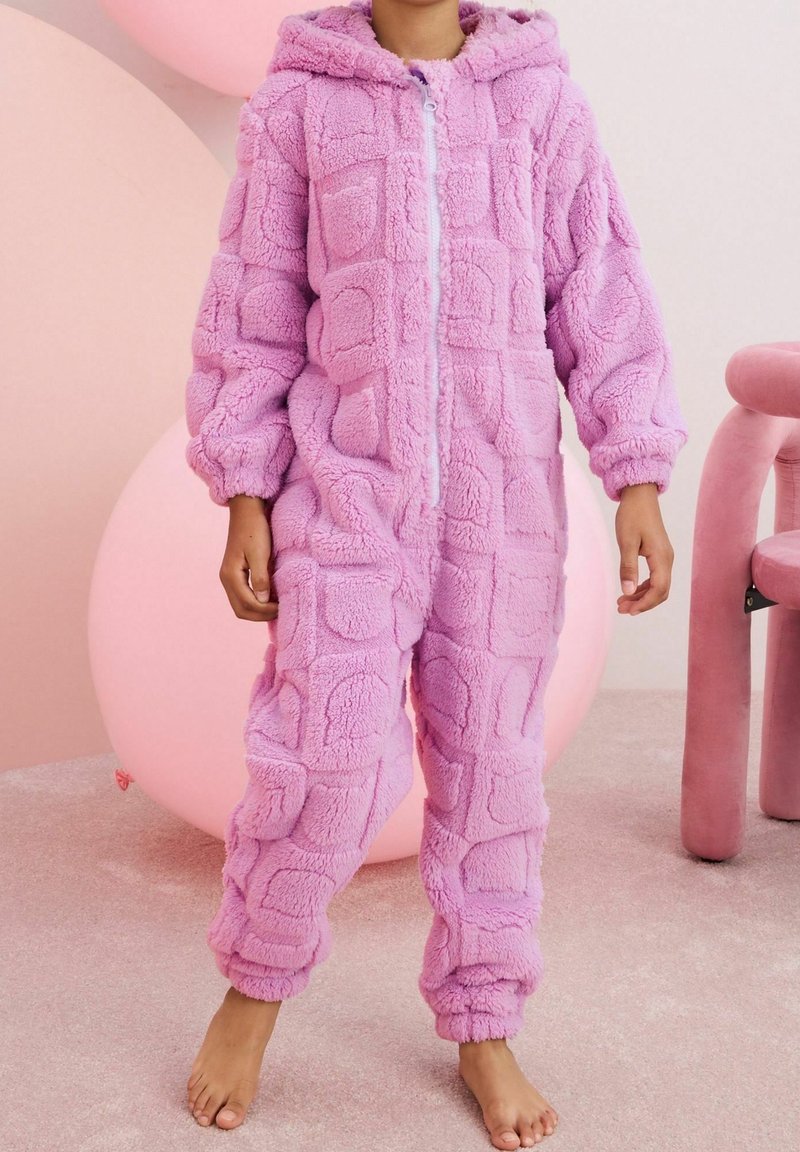 Enfant portant une grenouillère en polaire douce rose avec capuche et fermeture éclair frontale, debout pieds nus sur un tapis près d'un grand ballon rose et d'une chaise.