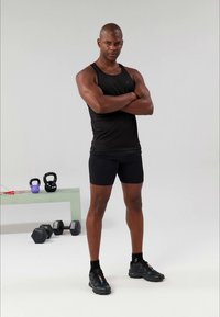 Camisola desportiva preta e calções, corpo musculoso, braços cruzados. Ténis pretos, equipamento de ginásio ao fundo, fundo cinza claro.