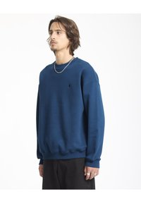 Marineblauwe sweatshirt in een ontspannen pasvorm, gemaakt van zachte stof met ribbels aan de uiteinden van de mouwen en de onderrand. Bevat een klein zwart logo op de borst.