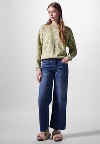 Vaaleanvihreä kuvioitu neule, siniset leveät farkut ja beige pörröinen tossut. Hiukset ovat suorat ja ulottuvat olkapäiden yli. Yksinkertainen, rento tyyli.