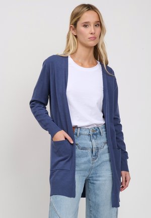 Frau trägt eine blaue offene Strickjacke über einem weißen T-Shirt und hellblauen hoch taillierten Jeans, stehend vor einem schlichten Hintergrund.