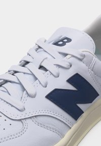 Λευκό δερμάτινο αθλητικό παπούτσι με μπλε λογότυπο "N" στο πλάι, λευκά κορδόνια δεμένα και λευκή σόλα με μπεζ λεπτομέρειες.