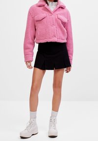 Giacca cropped rosa morbida con due tasche frontali, chiusura con bottoni, indossata sopra una gonna nera plissettata e sneakers bianche con zeppa e calzini grigi.