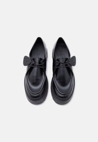 Ζεύγος μαύρων δερμάτινων μοκασίνων Hugo με στρογγυλεμένες μύτες, παχιά λαστιχένια σόλα και ετικέτες με το λογότυπο προσαρτημένες στα κορδόνια, απεικονιζόμενα από πάνω.