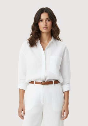 Vrouw met golvend bruin haar draagt een wit overhemd dat in een witte broek met een bruine riem is gestopt, staand tegen een effen achtergrond.