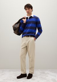 Junger Mann steht drinnen und trägt ein blau-navy gestreiftes Rugbyshirt, beige Hosen, dunkle Schuhe und hält eine kariertes Tasche über der Schulter.