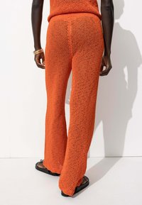 Un pantalon évasé tricoté orange présente une texture à mailles lâches. La taille est élastique et l'ourlet a un bord festonné. Des sandales plates noires sont portées.