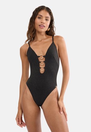 Frau trägt einen schwarzen Einteiler-Badeanzug mit dünnen Trägern und drei dekorativen goldenen Ringen entlang des tiefen V-Ausschnitts.