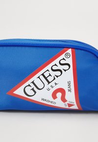 Sacca in poliestere blu con un logo triangolare rosso che presenta la scritta "GUESS" e "WASHED JEANS". Chiusura con zip superiore.