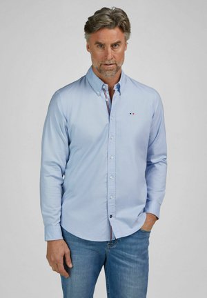 Man draagt lichtblauw overhemd met knopen en blauwe spijkerbroek, staat met één hand in zijn zak tegen een effen achtergrond.