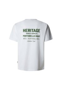 Balts kokvilnas T-krekls ar zaļu drukātu tekstu uz muguras, kas vēsta "HERITAGE GOODS & SUPPLIES PORTOBELLO ROAD 1973 - NOTTING HILL."