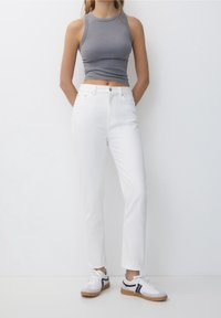 Gray ribbed tank top, white high-waisted jeans, and white athletic shoes with black accents. Model stands with hands in pockets.